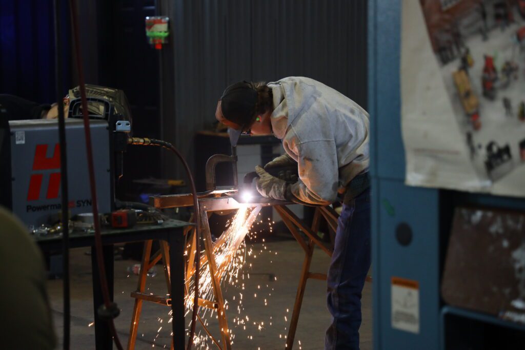 A student welder in protective gear works on a metal piece, producing a bright arc and cascade of sparks during BRTC's annual ARC welding competition.
