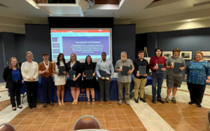A group of students and faculty pose together at BRTC's 2026 Phi Theta Kappa induction ceremony, holding certificate folders and lit candles, with a "Recognition of Inductees" slide projected behind them.