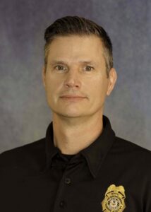 Professional headshot of a man with short dark hair in a black polo shirt with a gold badge on the chest.