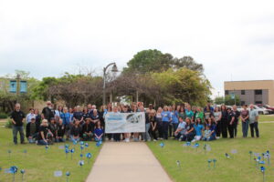 A large group of community members and BRTC staff pose outdoors on campus for the Child Abuse Awareness event, holding a "Pinwheels for Prevention" banner, surrounded by blue pinwheels displayed in the grass.