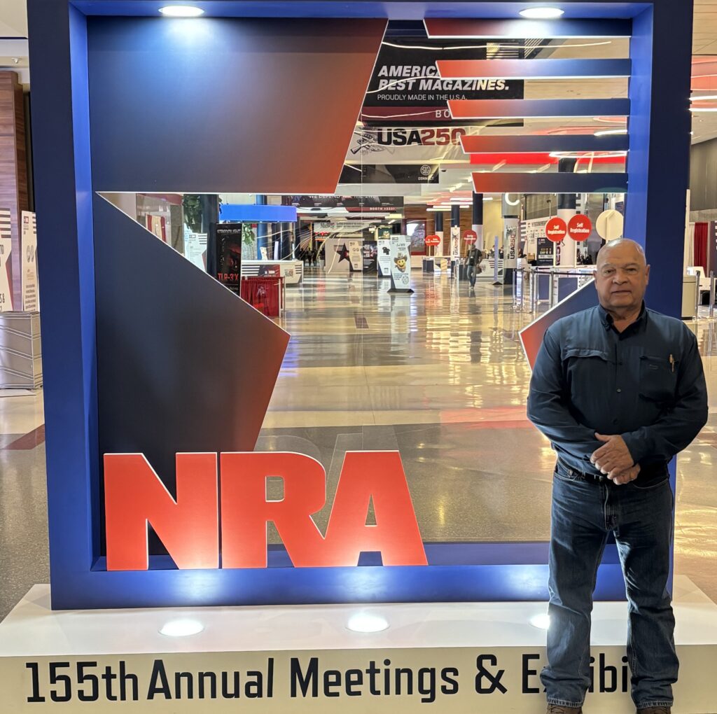 Gunsmithing Instructor Chuck Coe standing in front of an NRA sign at an NRA conference.