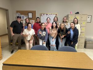 A group of students and an instructor stand in a classroom, each student holding a small pin. They are posing in front of a bulletin board, a calendar, and a set of lockers.