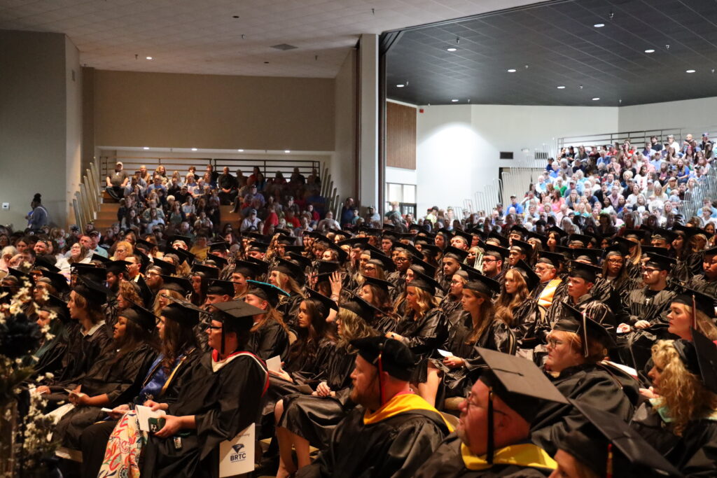 A large crowd of graduates wearing black caps and gowns sits in an auditorium during a commencement ceremony.
