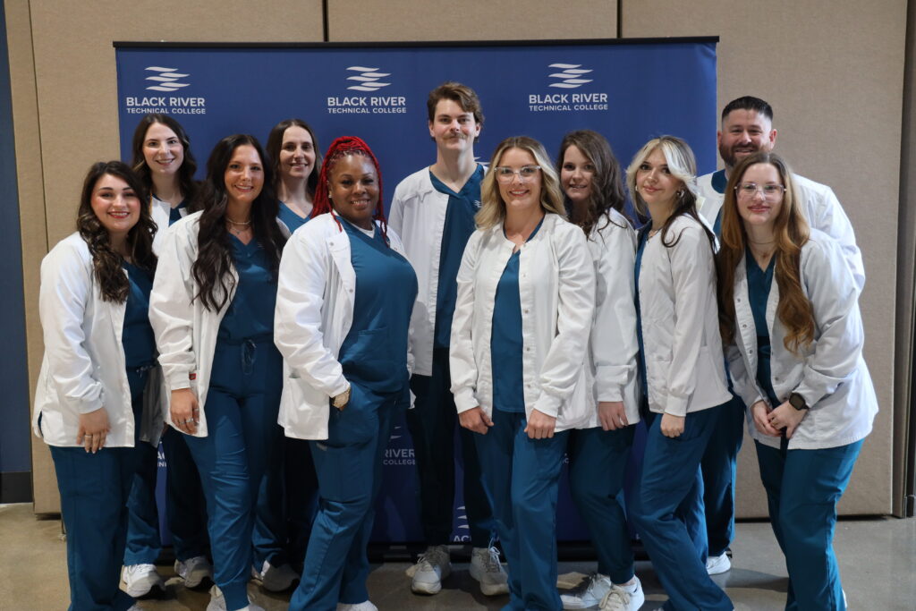 A group of eleven healthcare students pose together in front of a blue backdrop featuring the Black River Technical College logo.