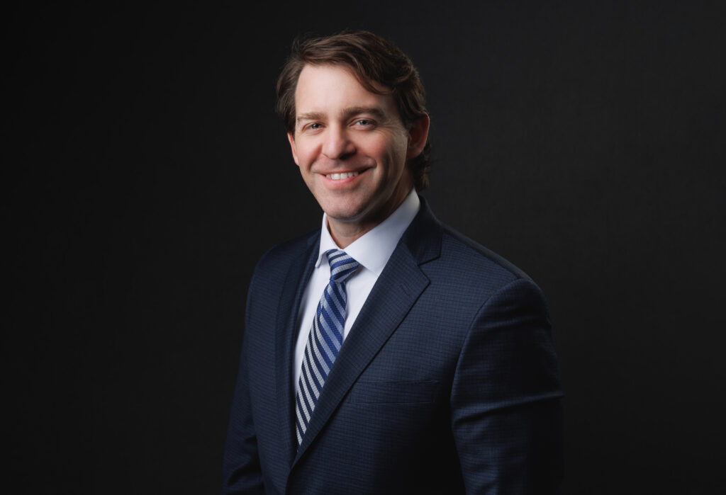 A professional studio headshot of a smiling man with short brown hair, captured from the chest up against a solid black background. He is wearing a dark blue textured suit jacket over a white dress shirt and a blue and white striped tie.