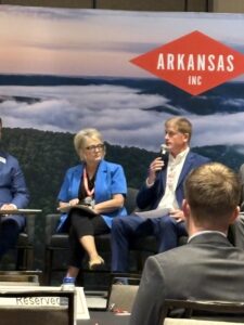 A panel discussion stage features a large backdrop of a foggy mountain landscape with a red diamond logo that reads "ARKANSAS INC". On stage, a woman with short blonde hair and glasses sits in a black chair, wearing a bright blue short-sleeved blazer over a black top and dark pants, holding a microphone and papers. To her right, a man in a blue suit and white dress shirt speaks into a handheld microphone while holding documents.
