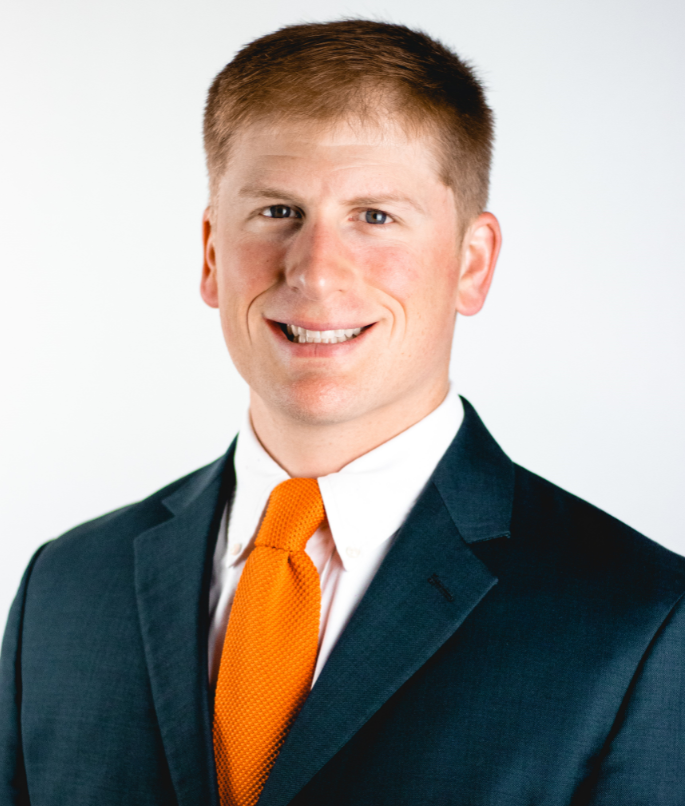 A professional studio headshot of a smiling man with short light brown hair, captured from the chest up against a plain white background. He is wearing a dark teal or charcoal suit jacket over a white collared dress shirt and a vibrant orange knit tie.