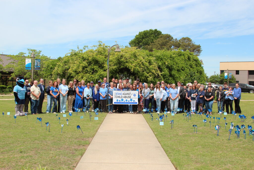 A large group of people stand together on a grassy lawn behind a banner that reads "Stand Against Child Abuse," which is decorated with blue pinwheels. Dozens of small blue pinwheels are also planted in the grass in front of the group, and a mascot in a blue bird costume stands to the far left.