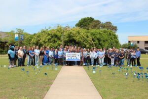 A large group of people stand together on a grassy lawn behind a banner that reads "Stand Against Child Abuse," which is decorated with blue pinwheels. Dozens of small blue pinwheels are also planted in the grass in front of the group, and a mascot in a blue bird costume stands to the far left.