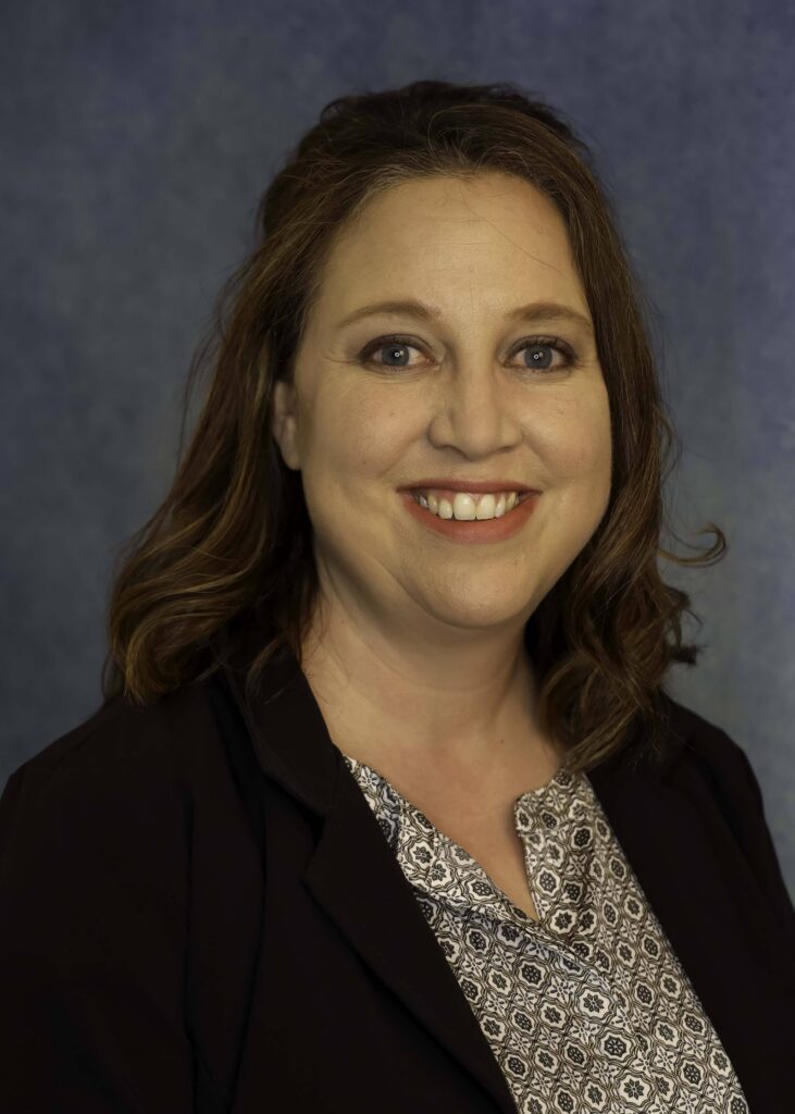 Woman with dark brown hair wearing a patterned shirt and blazer smiles for a headshot photo.
