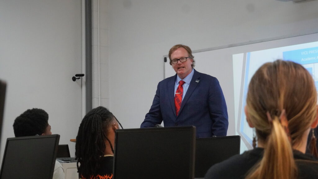 A man wearing a blue blazer and red tie presents to a classroom of students seated behind computer monitors.