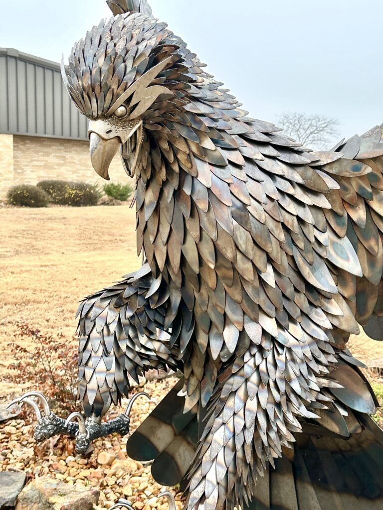 A close-up, side-profile view of a large eagle sculpture crafted entirely from welded, overlapping metal plates that resemble feathers.