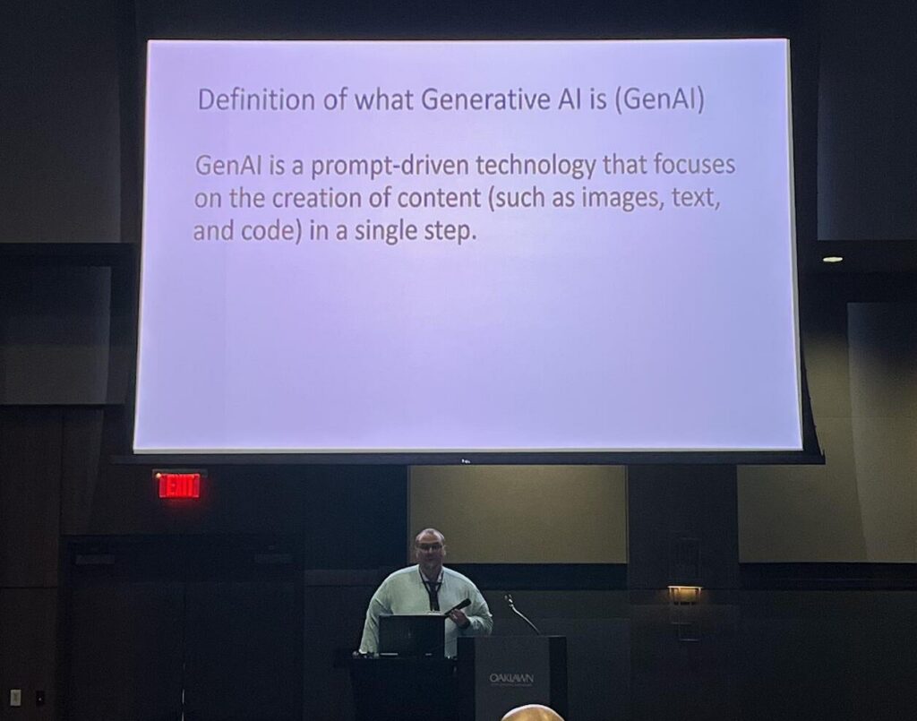 A man in glasses and a light-colored button up shirt stands behind a podium while speaking. A presentation slide defining Generative AI hangs behind him.