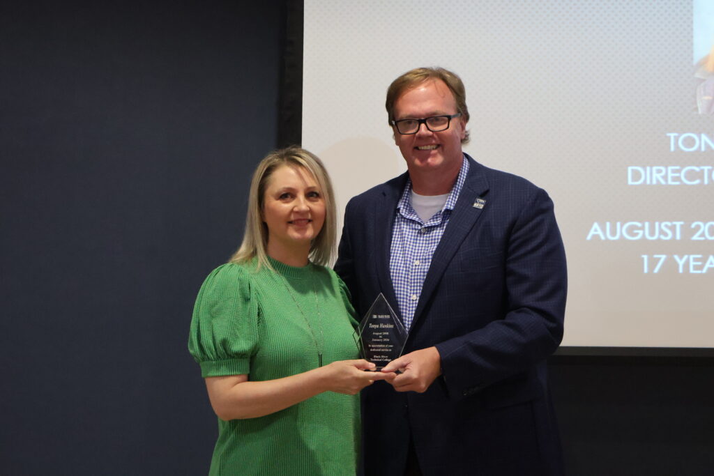 A medium shot shows a smiling woman with blonde, shoulder-length hair wearing a textured green dress while holding a clear, flame-shaped glass award alongside a tall, smiling man.