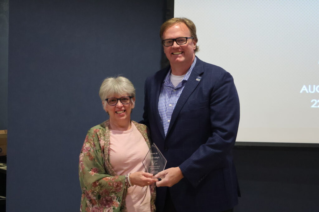 A medium shot shows a smiling woman with short silver hair and glasses receiving a clear, flame-shaped glass award from a tall, smiling man wearing glasses and a dark blue suit jacket over a checkered shirt.