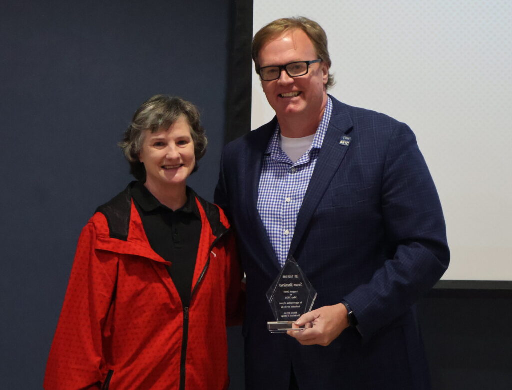A woman in a red jacket and a man in a navy blazer stand together smiling while holding a clear, diamond-shaped award.