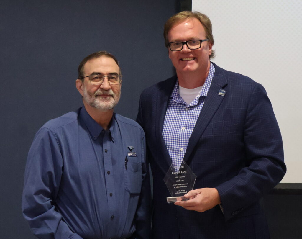 A man wearing a bule button-down shirt and glasses stands next to a man wearing a navy blue blazer, a blue checkered shirt, and glasses. The man in the checkered shirt is holding a small, clear award.