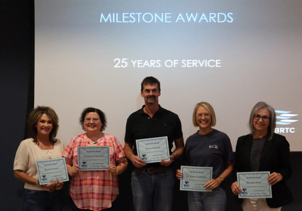 Five individuals stand in a row smiling while holding up paper certificates. A large projection screen behind them displays the text "MILESTONE AWARDS", "25 YEARS OF SERVICE", and a BRTC logo.