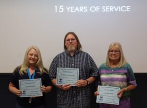 Three individuals stand side-by-side smiling in front of a projection screen that displays the text "15 YEARS OF SERVICE". Each person holds a BRTC "Milestone Award" certificate.