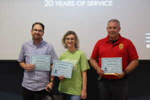 Three individuals stand side-by-side smiling in front of a projection screen displaying the text "20 YEARS OF SERVICE". Each person holds a BRTC "Milestone Award" certificate.