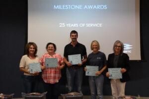 Five individuals stand in a row smiling while holding up paper certificates. A large projection screen behind them displays the text "MILESTONE AWARDS", "25 YEARS OF SERVICE", and a BRTC logo.