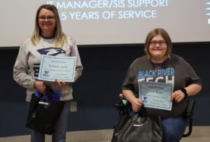 Two women smile while holding "Milestone Award" certificates in front of a projection screen. The woman on the left stands wearing a grey patterned sweatshirt and holds a certificate. The woman on the right sits in a motorized wheelchair wearing a dark grey shirt and holds a certificate.