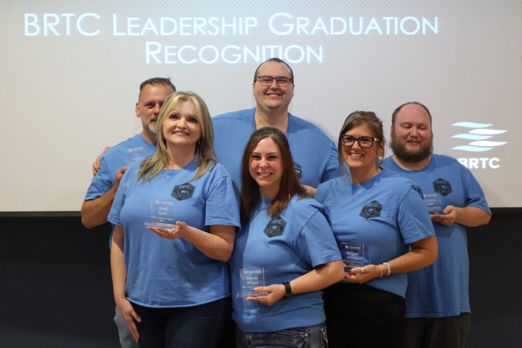 Six individuals wearing matching blue BRTC Leadership t-shirts pose holding awards at a recognition event.
