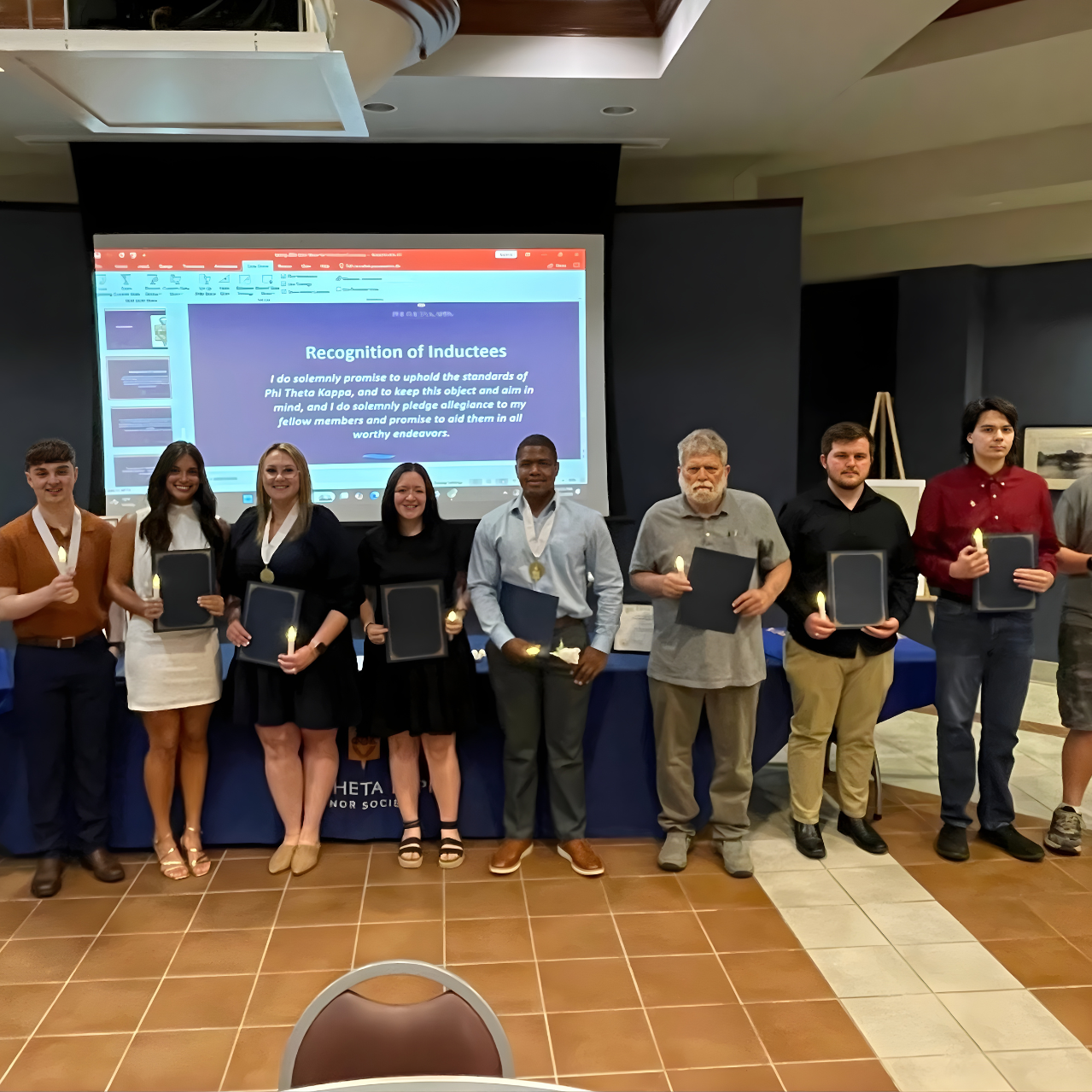 A group of students and faculty pose together at BRTC's 2026 Phi Theta Kappa induction ceremony, holding certificate folders and lit candles, with a "Recognition of Inductees" slide projected behind them.