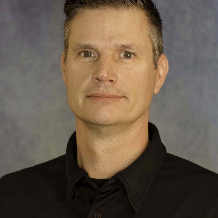 Professional headshot of a man with short dark hair in a black polo shirt with a gold badge on the chest.
