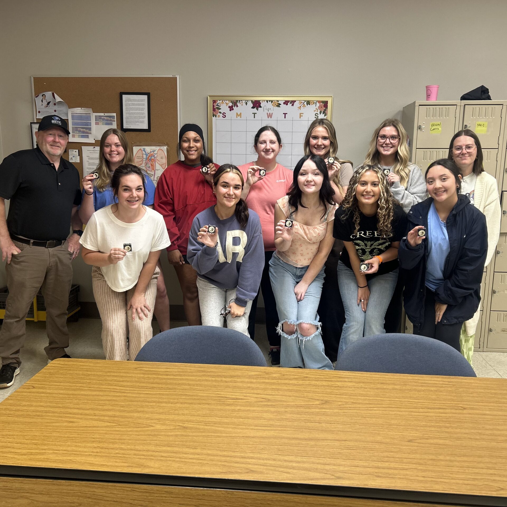 A group of students and an instructor stand in a classroom, each student holding a small pin. They are posing in front of a bulletin board, a calendar, and a set of lockers.