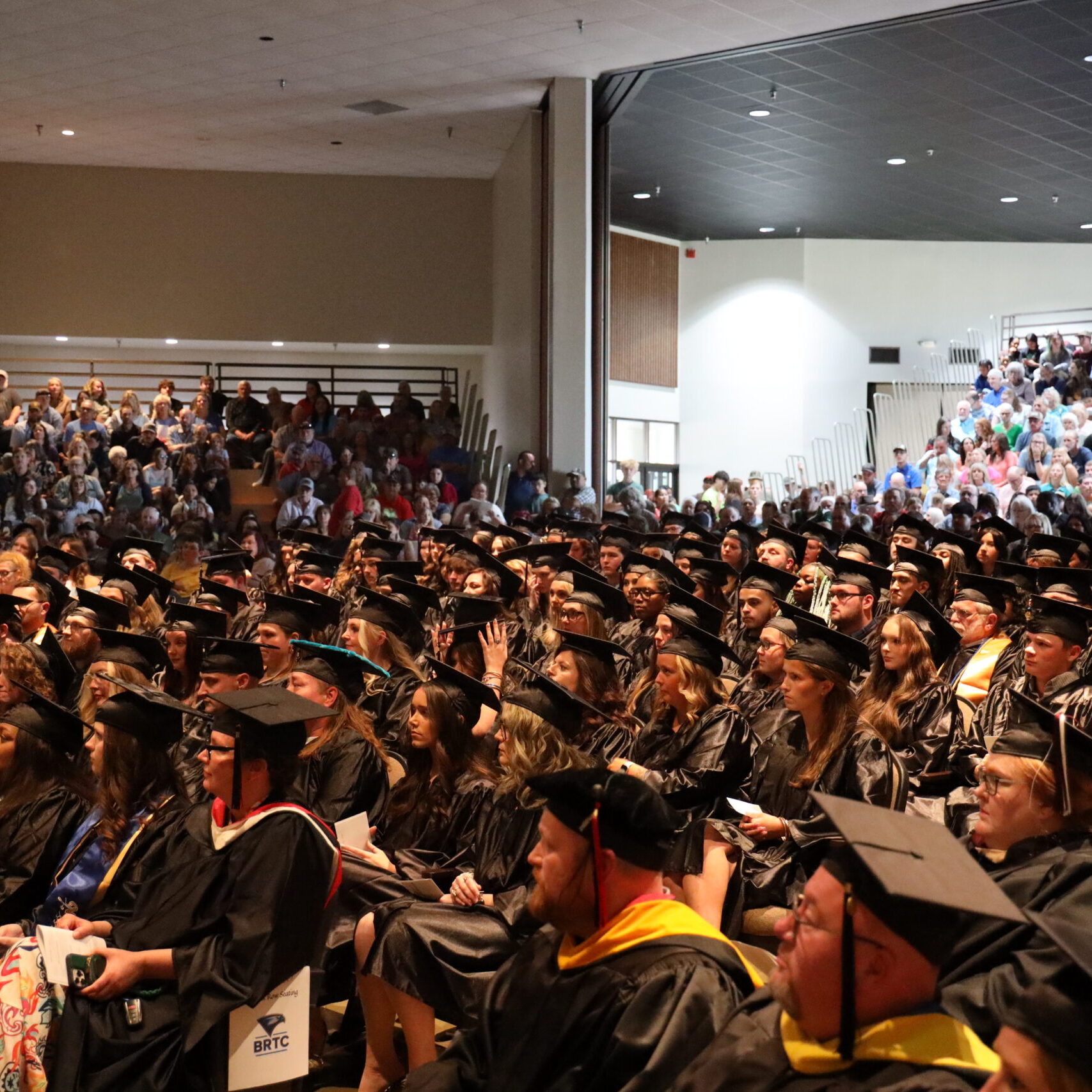 A large crowd of graduates wearing black caps and gowns sits in an auditorium during a commencement ceremony.