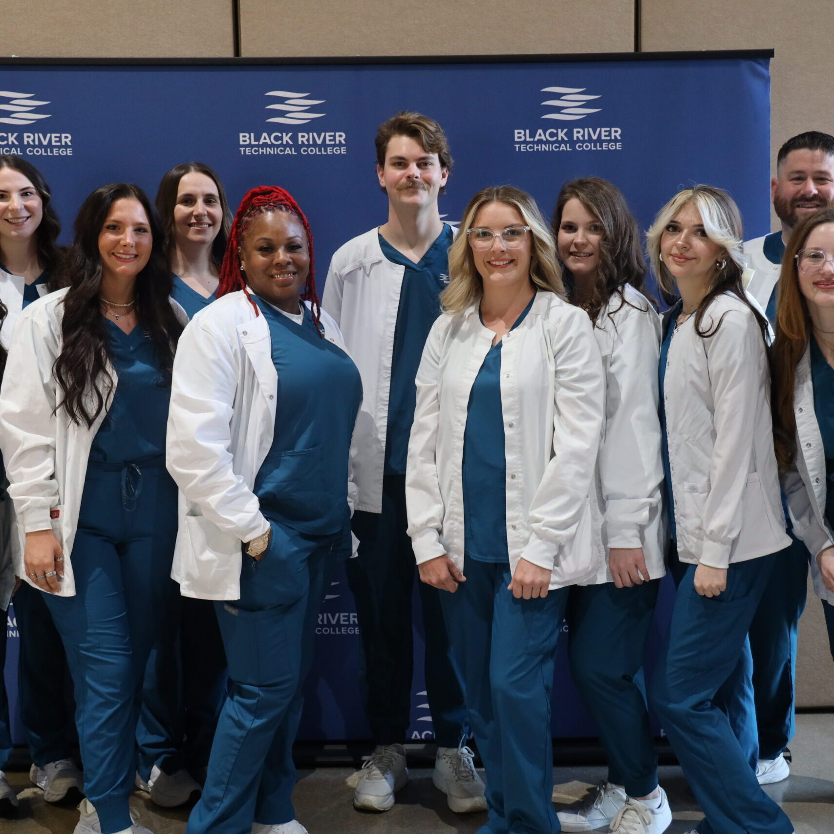 A group of eleven healthcare students pose together in front of a blue backdrop featuring the Black River Technical College logo.