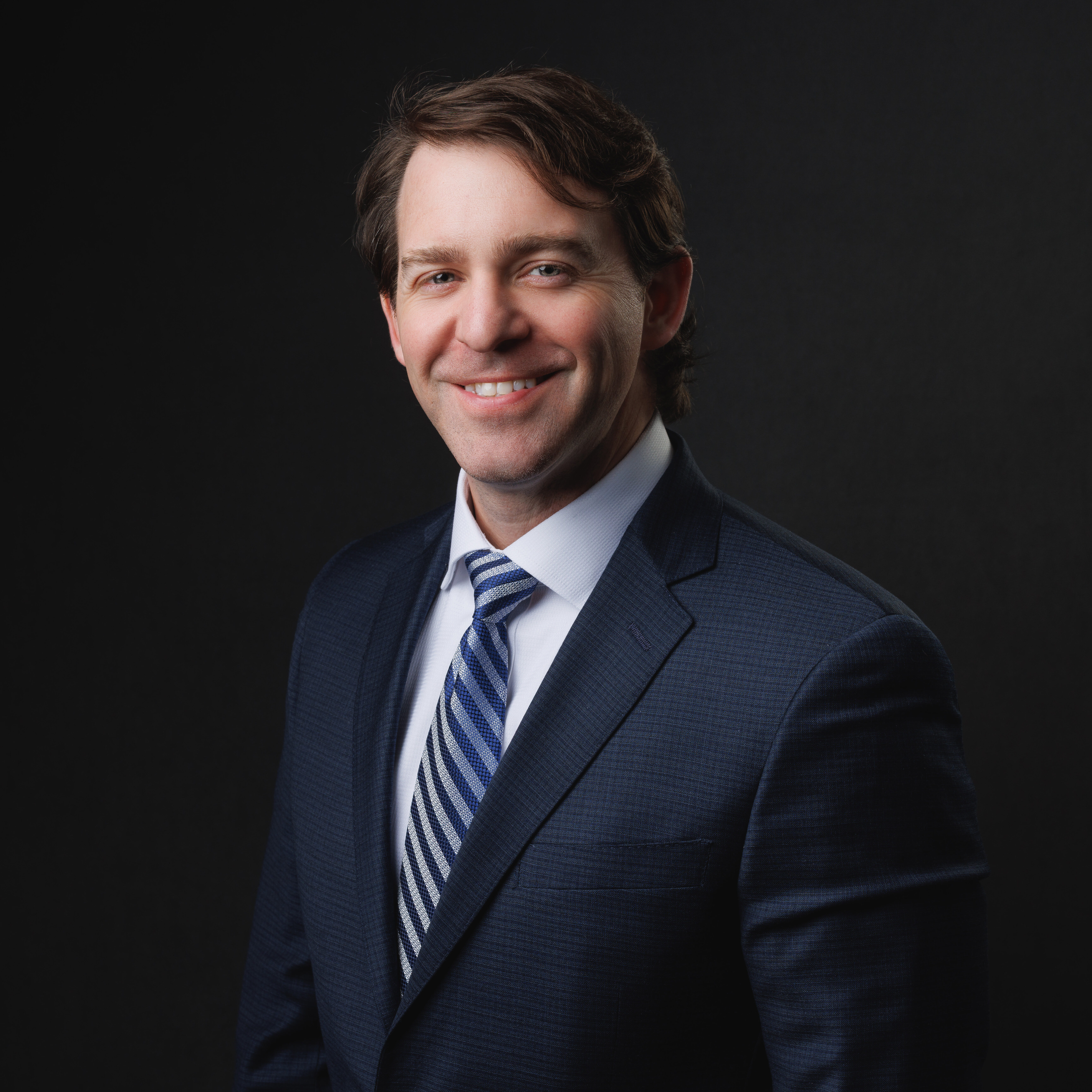 A professional studio headshot of a smiling man with short brown hair, captured from the chest up against a solid black background. He is wearing a dark blue textured suit jacket over a white dress shirt and a blue and white striped tie.
