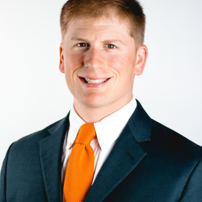 A professional studio headshot of a smiling man with short light brown hair, captured from the chest up against a plain white background. He is wearing a dark teal or charcoal suit jacket over a white collared dress shirt and a vibrant orange knit tie.