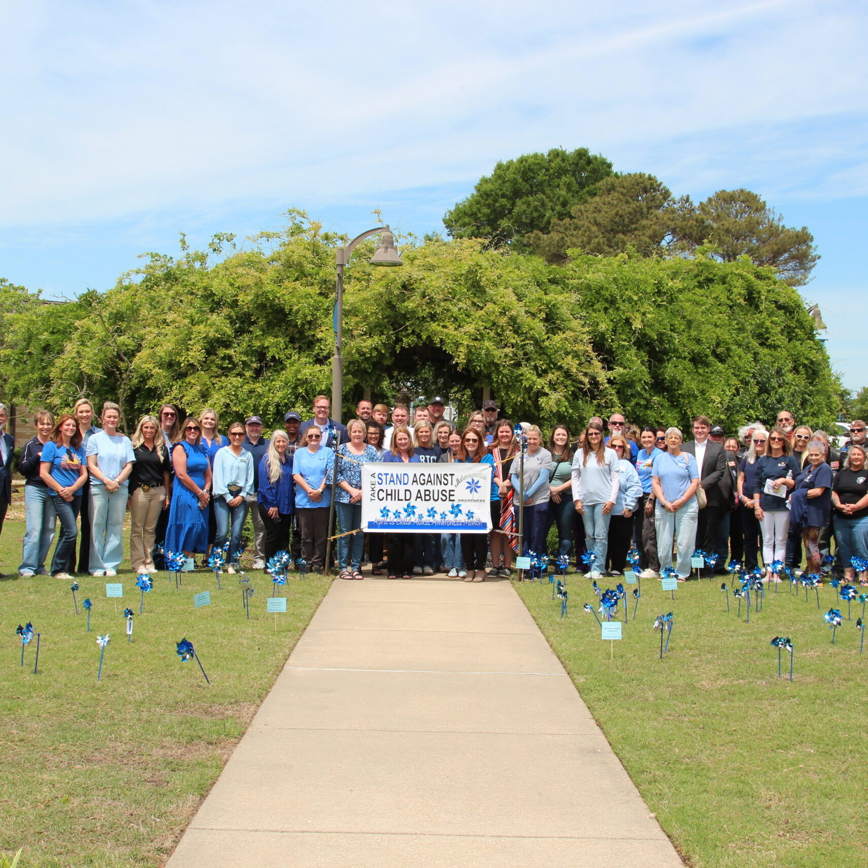 A large group of people stand together on a grassy lawn behind a banner that reads "Stand Against Child Abuse," which is decorated with blue pinwheels. Dozens of small blue pinwheels are also planted in the grass in front of the group, and a mascot in a blue bird costume stands to the far left.