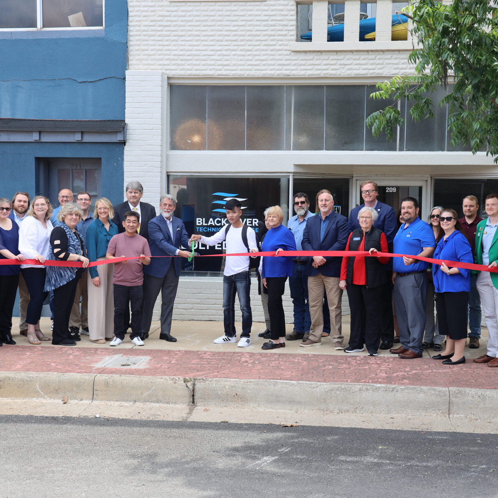 A group of community leaders and representatives hold a long red ribbon for a ceremonial cutting in front of the Black River Technical College Adult Education building in downtown Paragould.