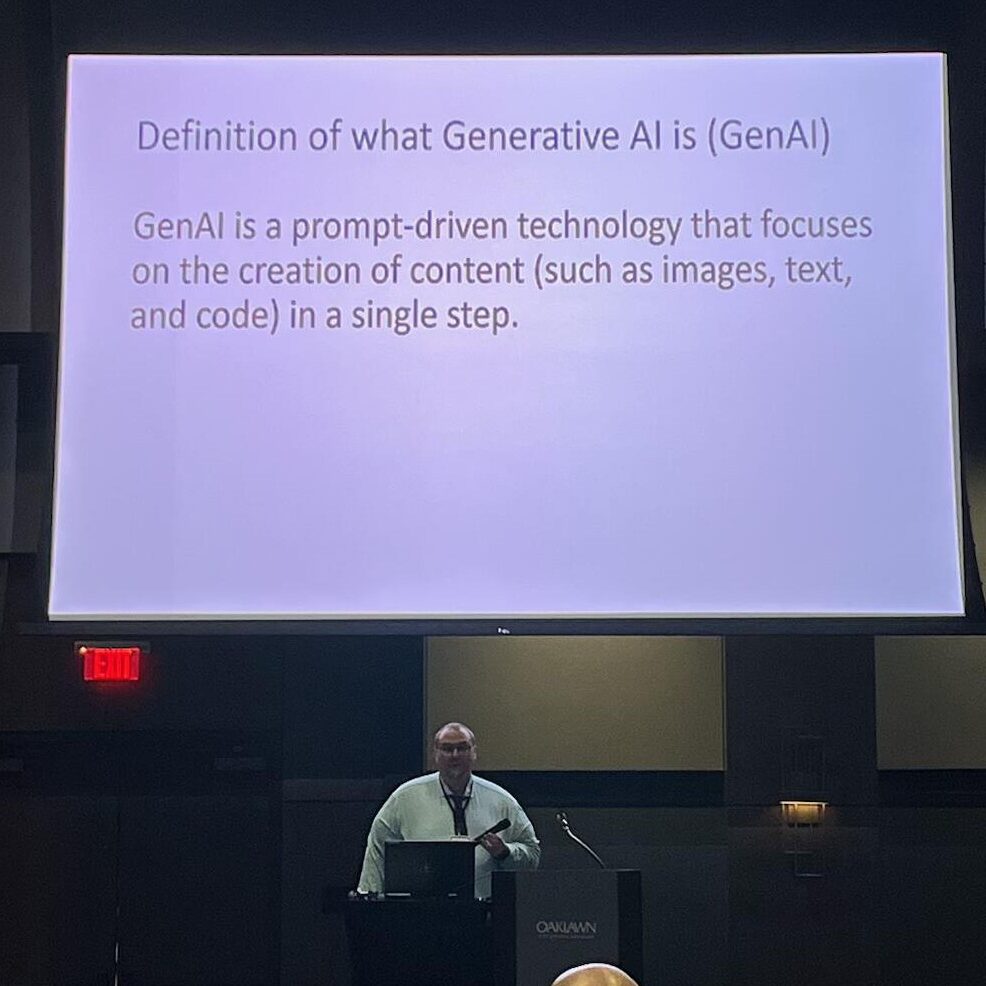 A man in glasses and a light-colored button up shirt stands behind a podium while speaking. A presentation slide defining Generative AI hangs behind him.