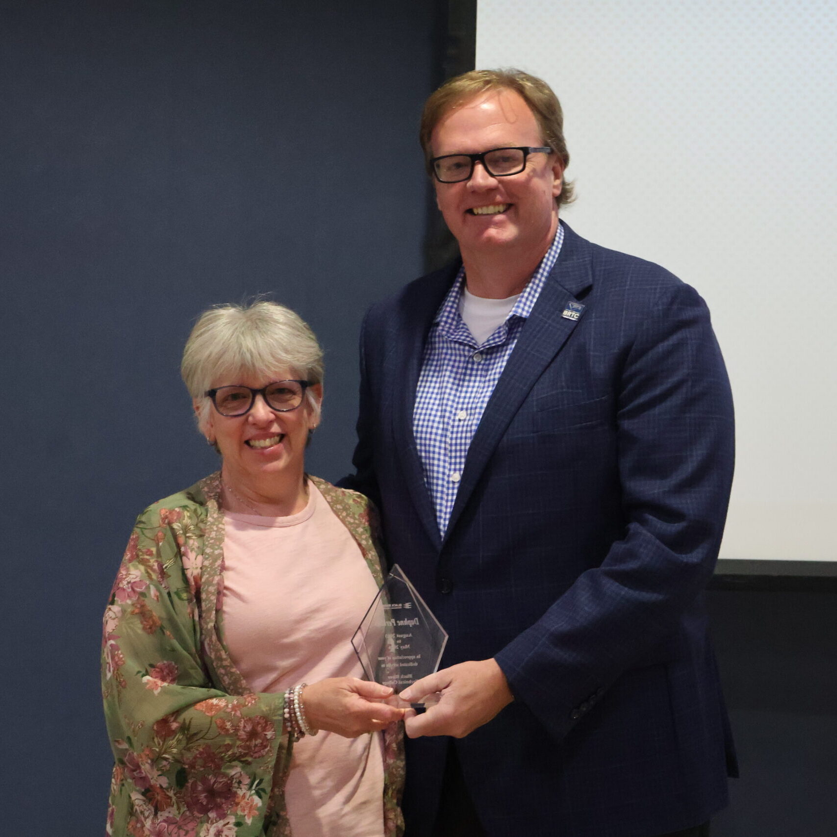 A medium shot shows a smiling woman with short silver hair and glasses receiving a clear, flame-shaped glass award from a tall, smiling man wearing glasses and a dark blue suit jacket over a checkered shirt.