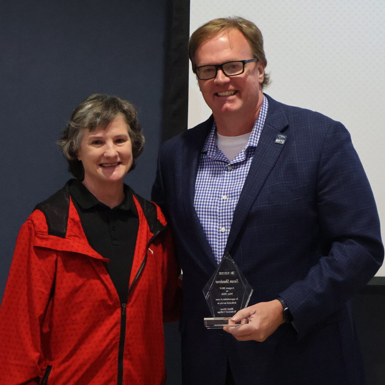 A woman in a red jacket and a man in a navy blazer stand together smiling while holding a clear, diamond-shaped award.