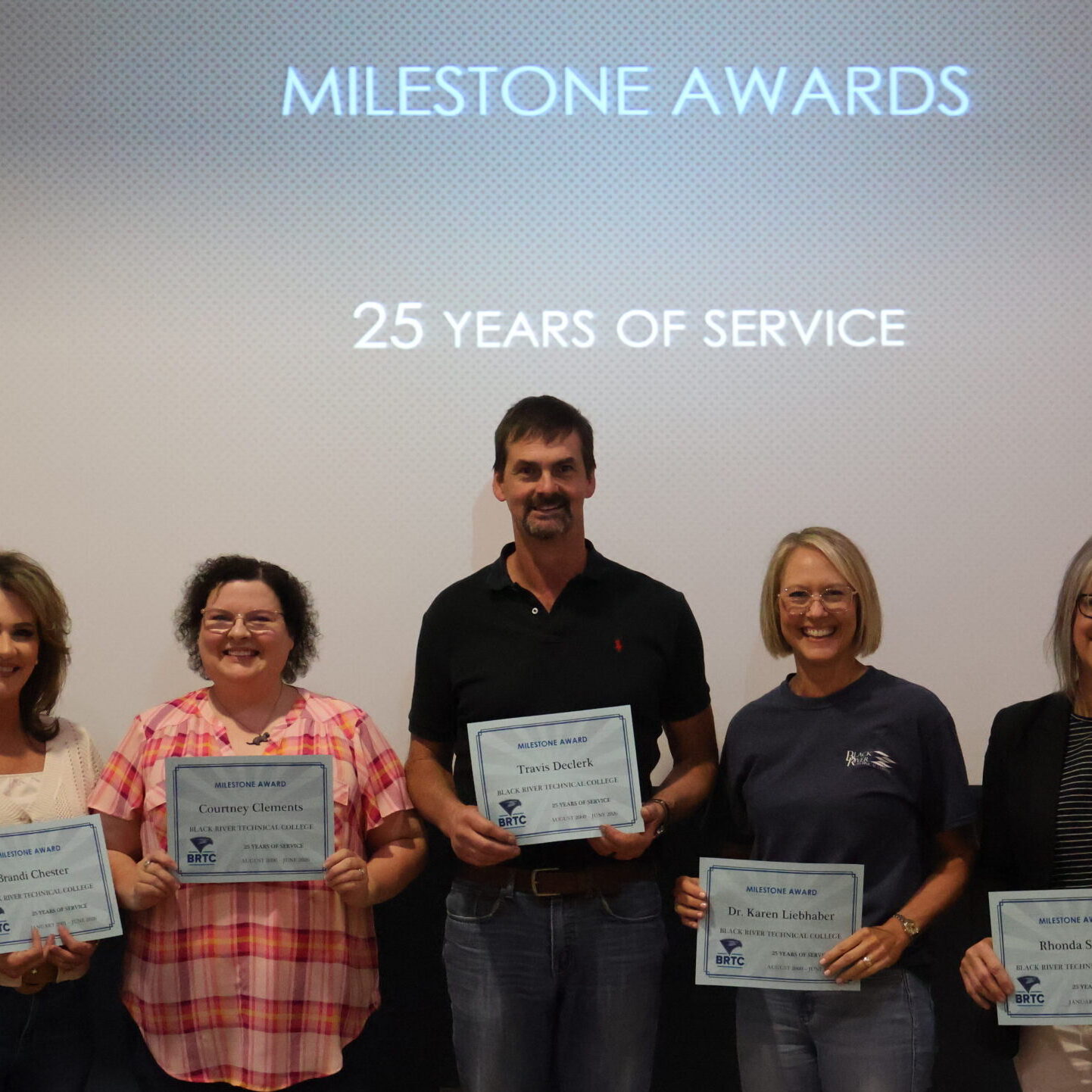 Five individuals stand in a row smiling while holding up paper certificates. A large projection screen behind them displays the text "MILESTONE AWARDS", "25 YEARS OF SERVICE", and a BRTC logo.