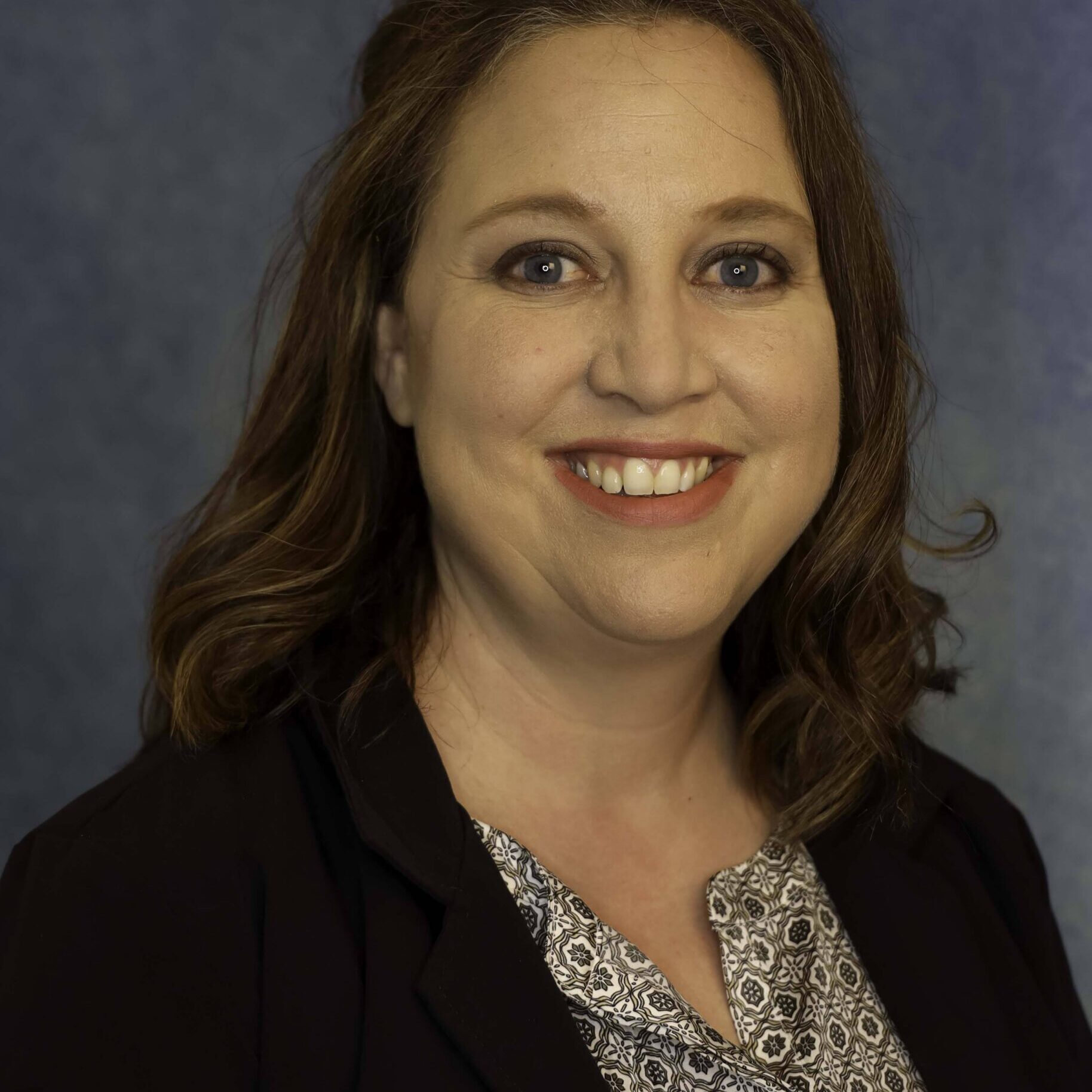 Woman with dark brown hair wearing a patterned shirt and blazer smiles for a headshot photo.