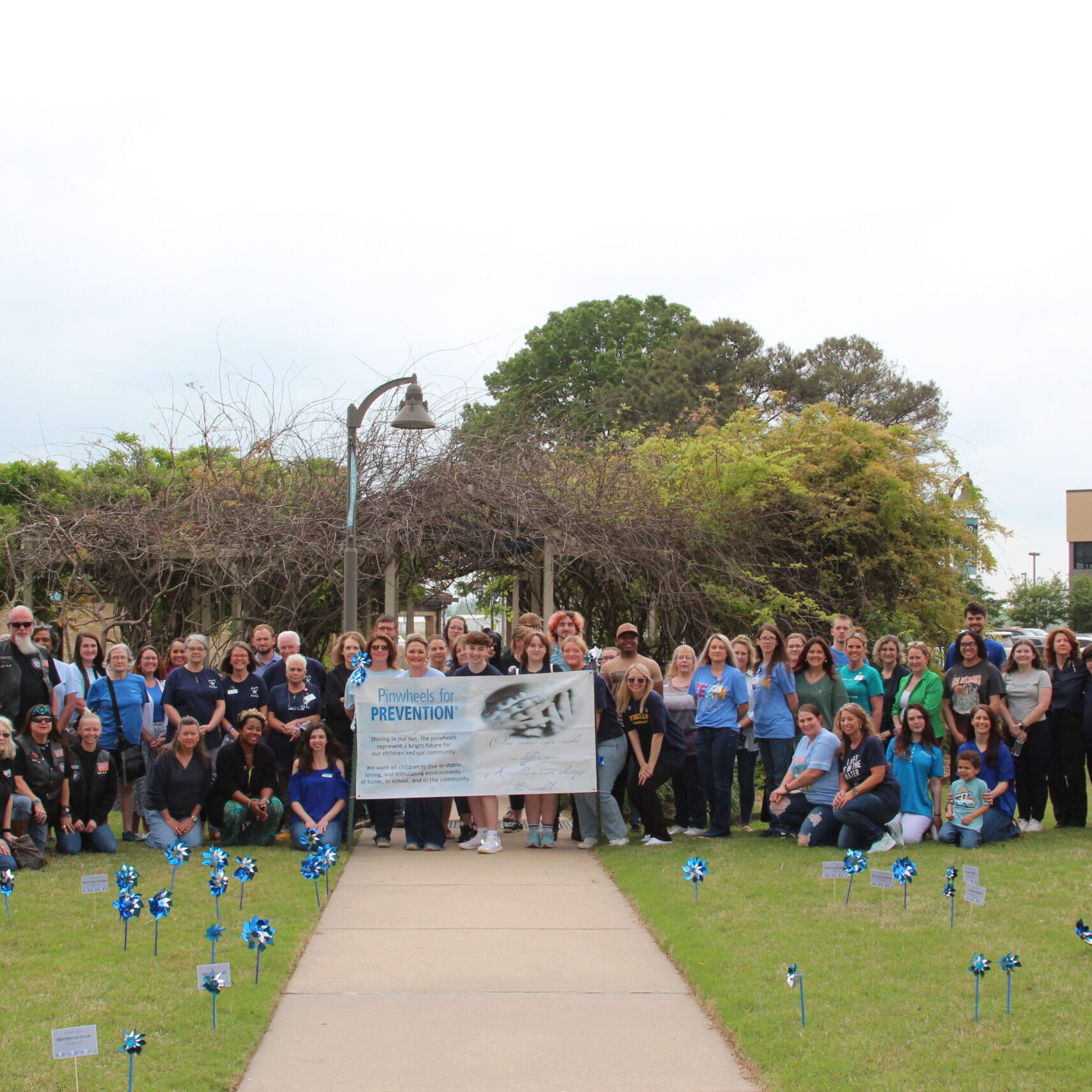A large group of community members and BRTC staff pose outdoors on campus for the Child Abuse Awareness event, holding a "Pinwheels for Prevention" banner, surrounded by blue pinwheels displayed in the grass.