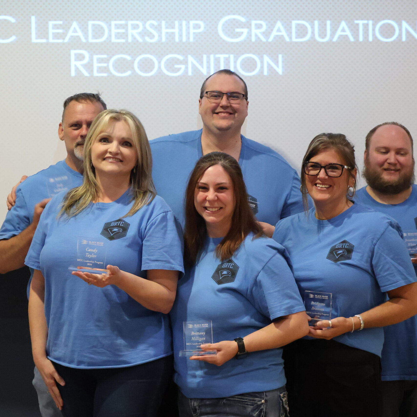 Six individuals wearing matching blue BRTC Leadership t-shirts pose holding awards at a recognition event.
