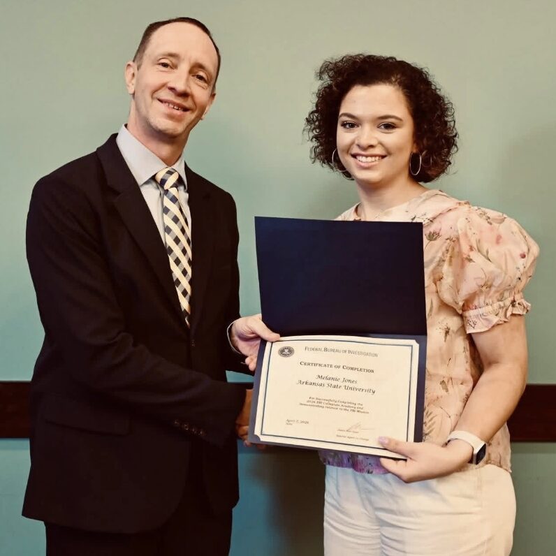 A young woman receives an FBI Certificate of Completion from a suited official, recognizing her successful completion of the FBI Collegiate Academy, representing Arkansas State University.
