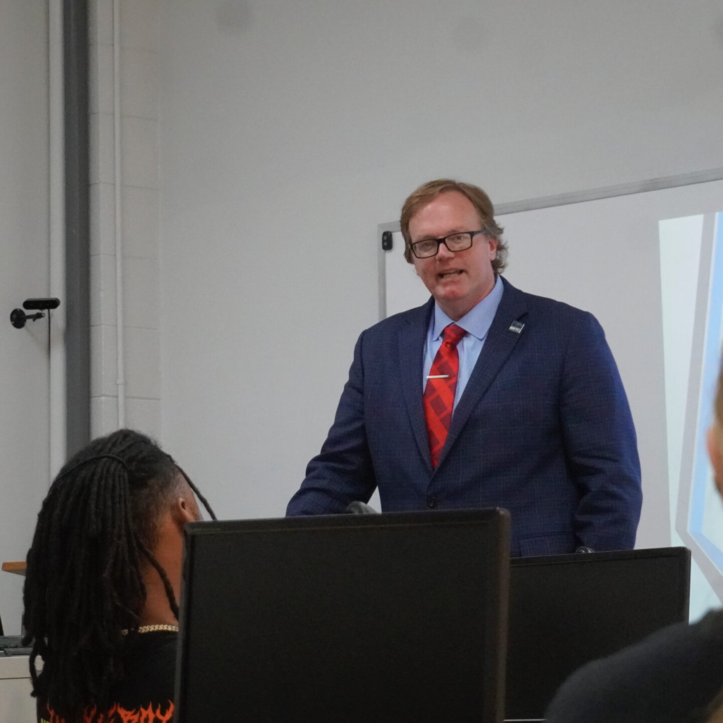 A man wearing a blue blazer and red tie presents to a classroom of students seated behind computer monitors.